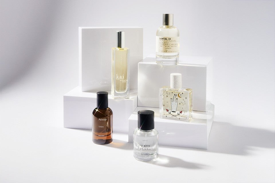 Five perfume bottles are arranged on white geometric blocks. The bottles vary in shape and color: one is tall and rectangular with a silver cap and light yellow liquid labeled "kai," another is round with a silver cap and clear liquid labeled "SANTAL 33," a third is square with a white cap and decorative design featuring a hand, moon, and stars, a fourth is amber-colored with a black cap labeled "Aesop," and the fifth is clear with a black cap labeled "DIRTY COCONUT." The background is a clean, light gray.