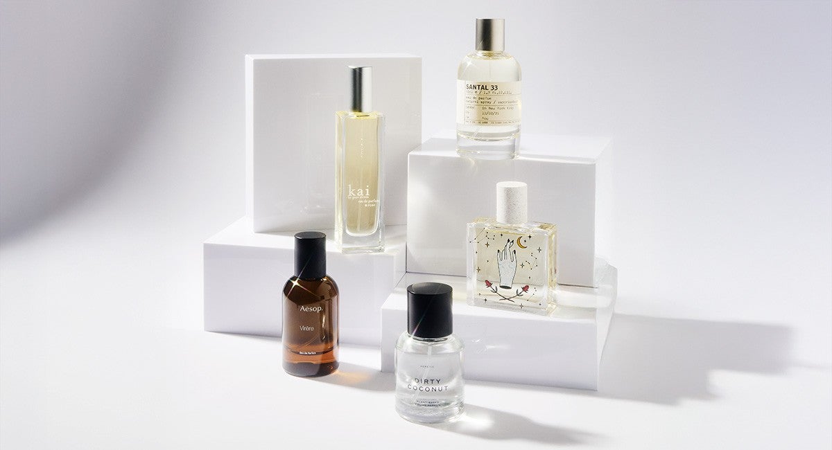 Five perfume bottles are arranged on white geometric blocks. The bottles vary in shape and color: one is tall and rectangular with a silver cap labeled "kai," another is round and clear with a black cap labeled "DIRTY COCONUT," a third is amber-colored with a black cap labeled "Aesop," a fourth is clear with a white cap featuring an illustration of a hand, crescent moon, and stars, and the fifth is clear with a silver cap labeled "SANTAL 33." The background is plain white with soft shadows.