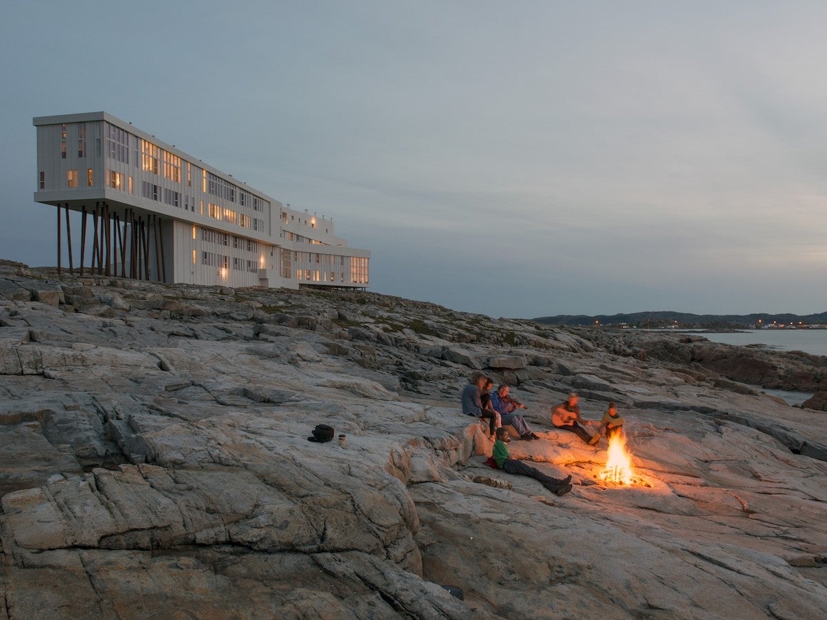 A modern, white building elevated on stilts sits on rocky terrain near the coast at dusk. In the foreground, a group of people are gathered around a bright campfire on the rocks, with one person playing a guitar. The sky is clear with soft evening light.
