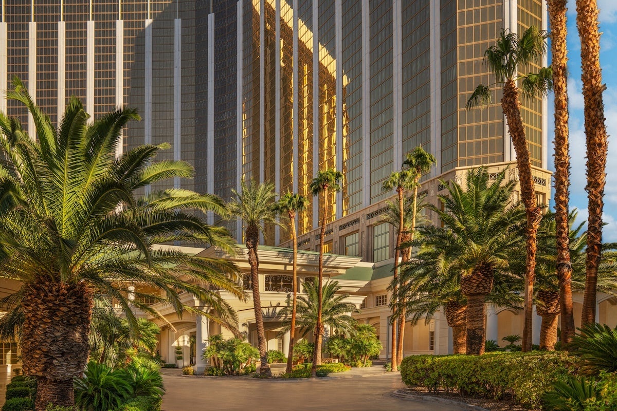 Tall palm trees and lush greenery surround the entrance of a large building with reflective golden windows, bathed in warm sunlight. The building features a covered driveway with white columns and a green roof.