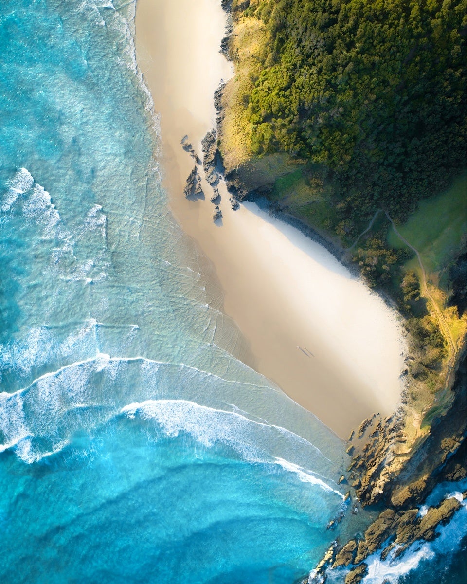 Aerial view of a sandy beach bordered by clear blue ocean waves on the left and dense green forest on the right. Rocky formations are visible along the shoreline, and a small path runs through the forest near the beach. The sunlight casts shadows on the forest and beach area.