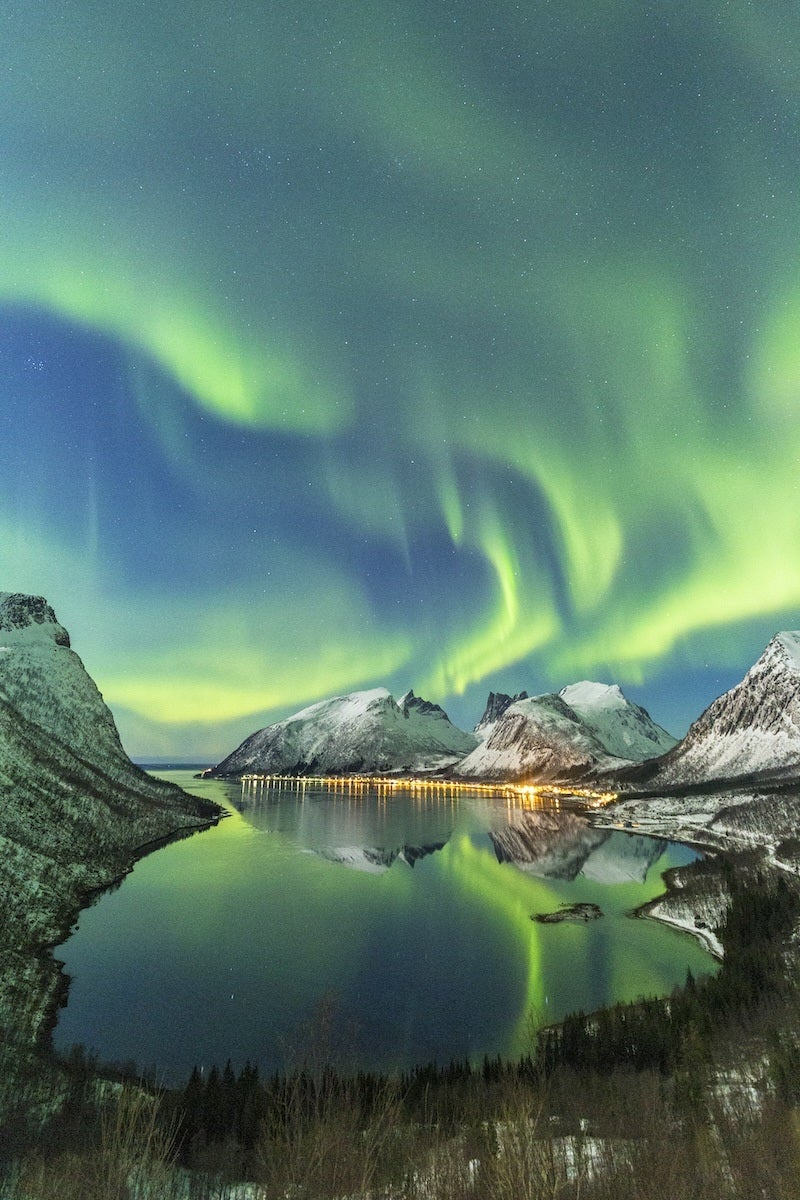 Northern lights glowing green and blue over snow-covered mountains, reflected in a calm fjord with a small illuminated settlement along the shoreline.