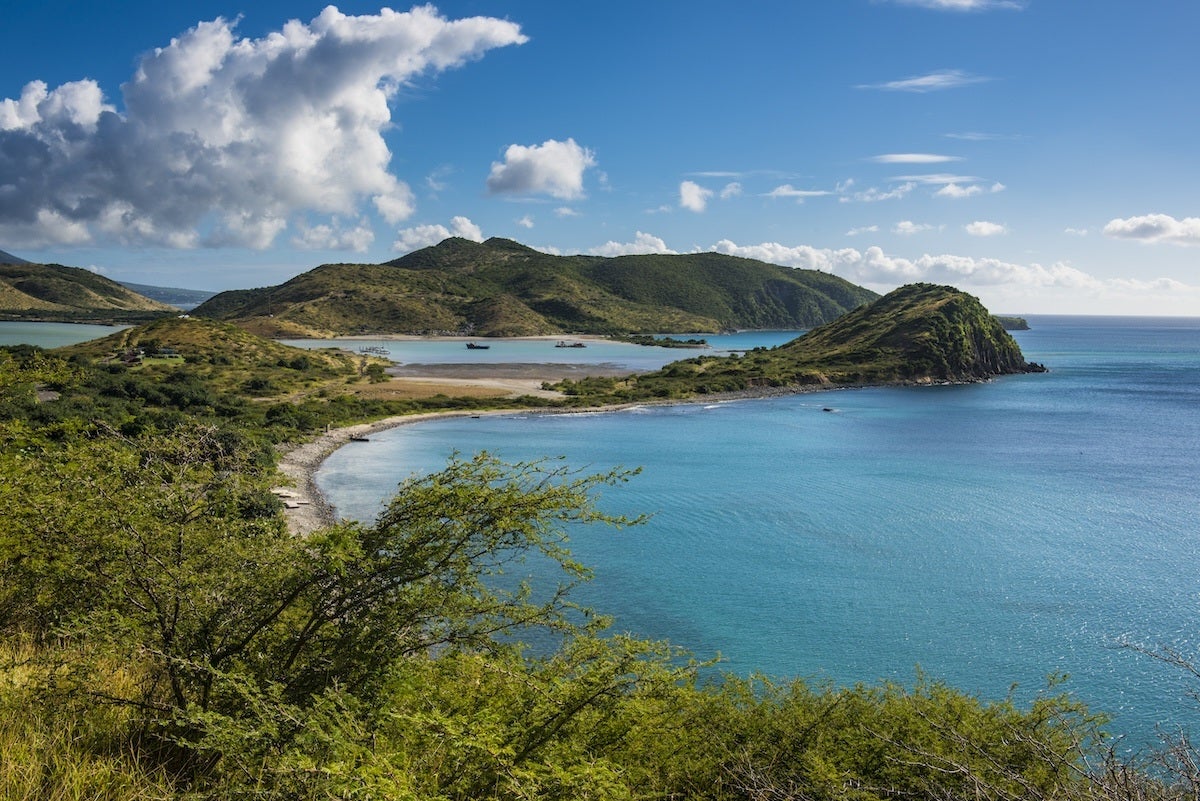 A coastal landscape featuring a curved shoreline with clear turquoise water. The foreground has green shrubs and vegetation, while the background shows rolling hills covered in greenery under a partly cloudy blue sky.