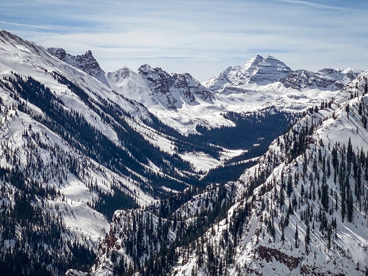 Snow-covered mountain range with dense clusters of evergreen trees scattered along the slopes under a partly cloudy sky.