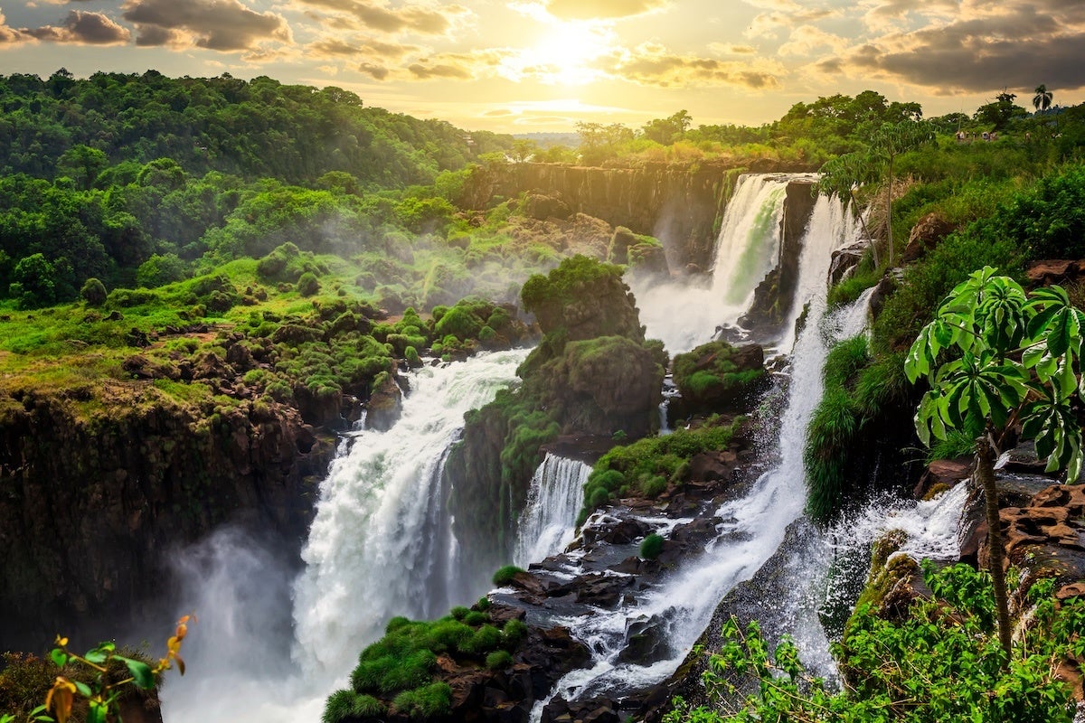 A scenic view of a large waterfall cascading over rocky cliffs surrounded by lush green vegetation under a partly cloudy sky with the sun setting or rising in the background. Mist rises from the base of the waterfall, and the landscape features dense forested hills.