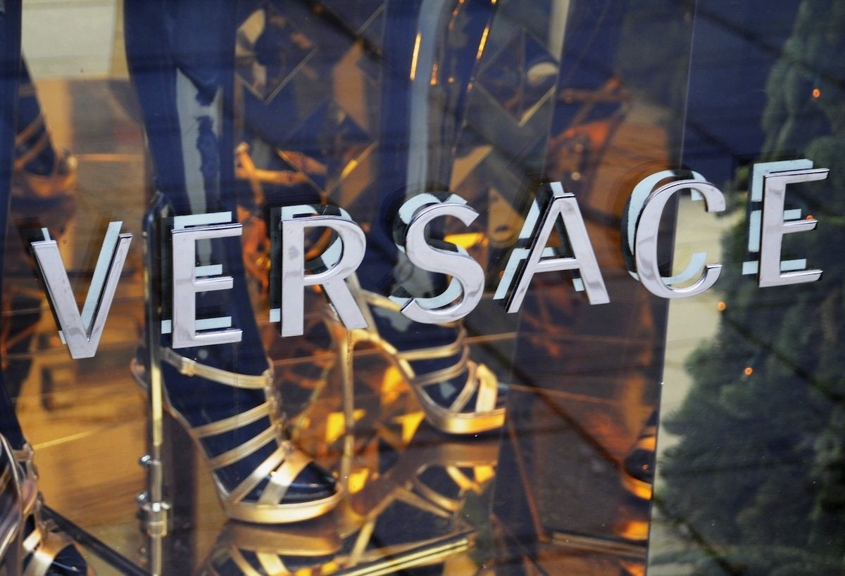 Versace logo displayed on a glass surface with a reflection of gold high-heeled shoes and a person wearing dark pants in the background.