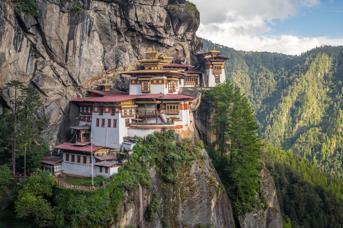 A traditional Bhutanese monastery built on the edge of a steep cliff, featuring white walls, red roofs, and ornate golden details. The structure is surrounded by lush green trees and dense forested mountains under a partly cloudy sky.
