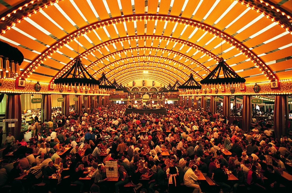 A large, crowded beer hall with long wooden tables filled with people. The hall has a high, arched ceiling adorned with rows of warm yellow lights and large, circular chandeliers. The atmosphere is lively and festive, with many people enjoying drinks and socializing. The walls are decorated with banners and drapes, contributing to a traditional, celebratory ambiance.