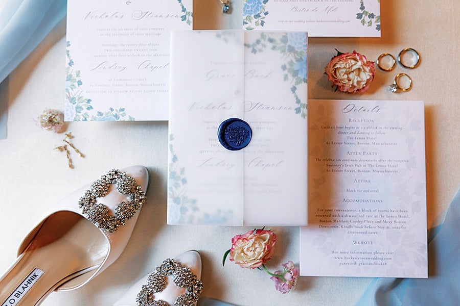 A pair of elegant white satin shoes with large, ornate rhinestone buckles are placed next to a set of wedding invitations and details cards. The invitations feature a floral blue and green design with cursive text. A translucent vellum envelope with a blue wax seal is centered among the stationery. There are also a few small flowers and three rings, including a diamond engagement ring, arranged nearby on a light-colored surface.