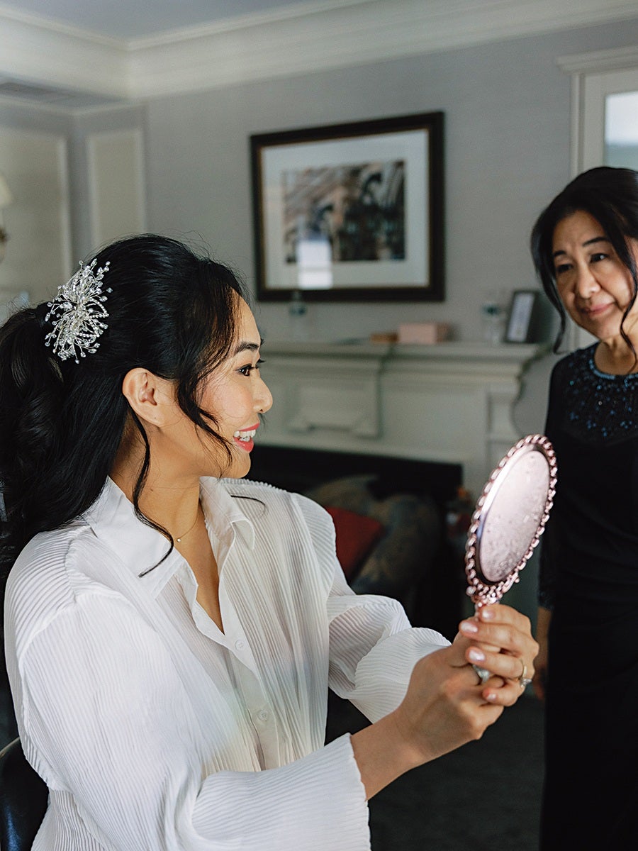A woman with long black hair styled with a decorative hair accessory is smiling and holding a pink ornate hand mirror. She is wearing a white, pleated blouse. Another woman, dressed in black, is standing nearby, looking at her with a gentle expression. The background features a framed picture on a light-colored wall and a fireplace.