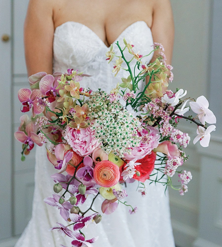 A bridal bouquet featuring a variety of flowers including pink and purple orchids, white and pink peonies, small white blossoms with green centers, and coral ranunculus, held by a bride wearing a strapless white lace wedding dress.