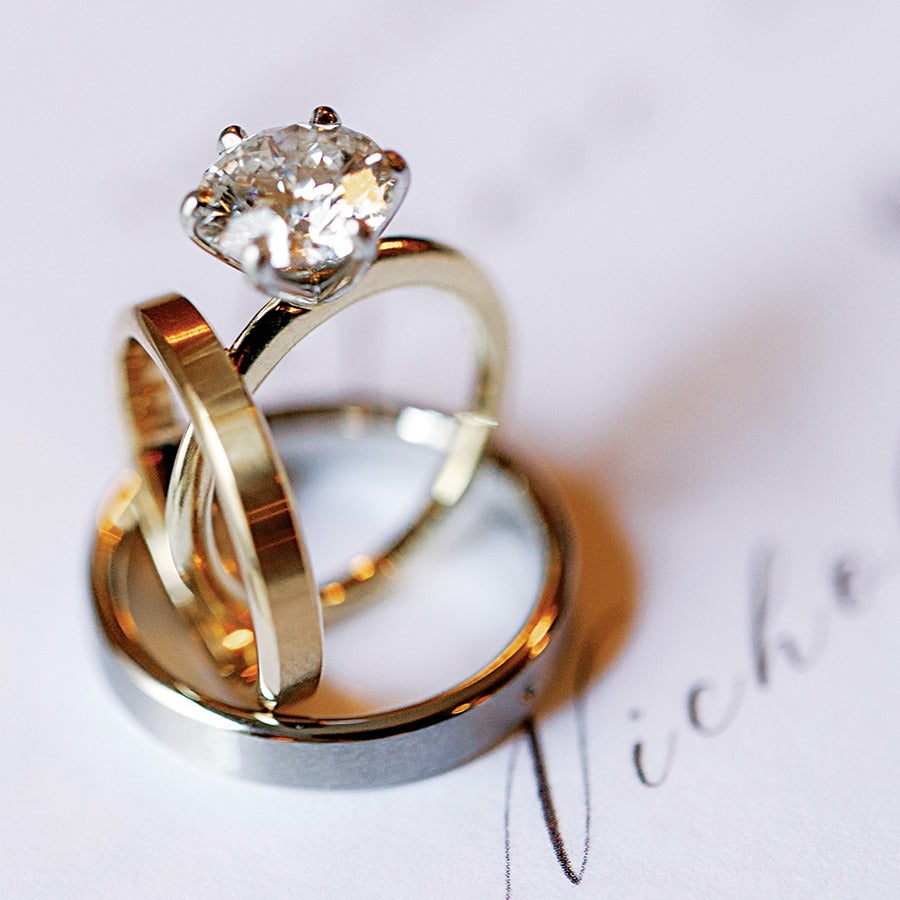 Three rings stacked together: a gold engagement ring with a large round diamond, a plain gold wedding band, and a wider silver or platinum wedding band, placed on a white surface with partially visible cursive writing.