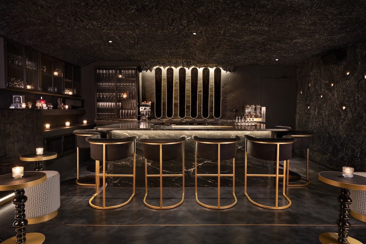 A modern, dimly lit bar with a dark textured ceiling and walls. The bar counter is made of dark marble with subtle lighting underneath. Five sleek bar stools with gold frames and black cushioned seats are lined up in front of the counter. Behind the bar, there are tall, narrow mirrors with backlighting and shelves filled with glassware. Small candles are placed on side tables and shelves, adding to the intimate ambiance.