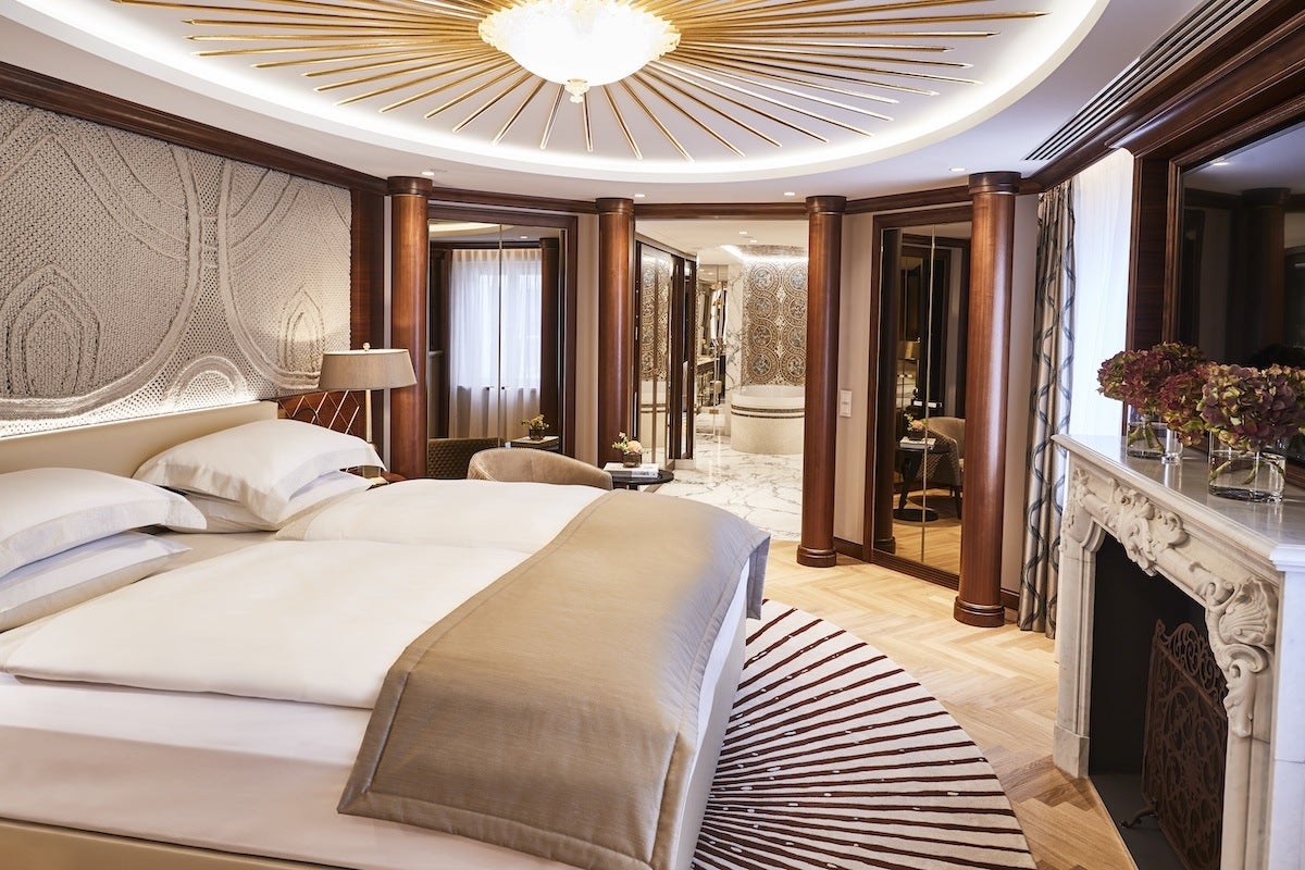 Luxurious bedroom with a large bed featuring white linens and a beige throw. The room has wooden columns, a decorative beige wall panel behind the bed, and a round rug with a radial pattern. A marble fireplace with floral arrangements sits to the right, and a large mirror above it reflects the room. In the background, a bathroom with ornate tile work and a bathtub is visible through an open doorway. The ceiling has a circular light fixture with gold rays extending outward.