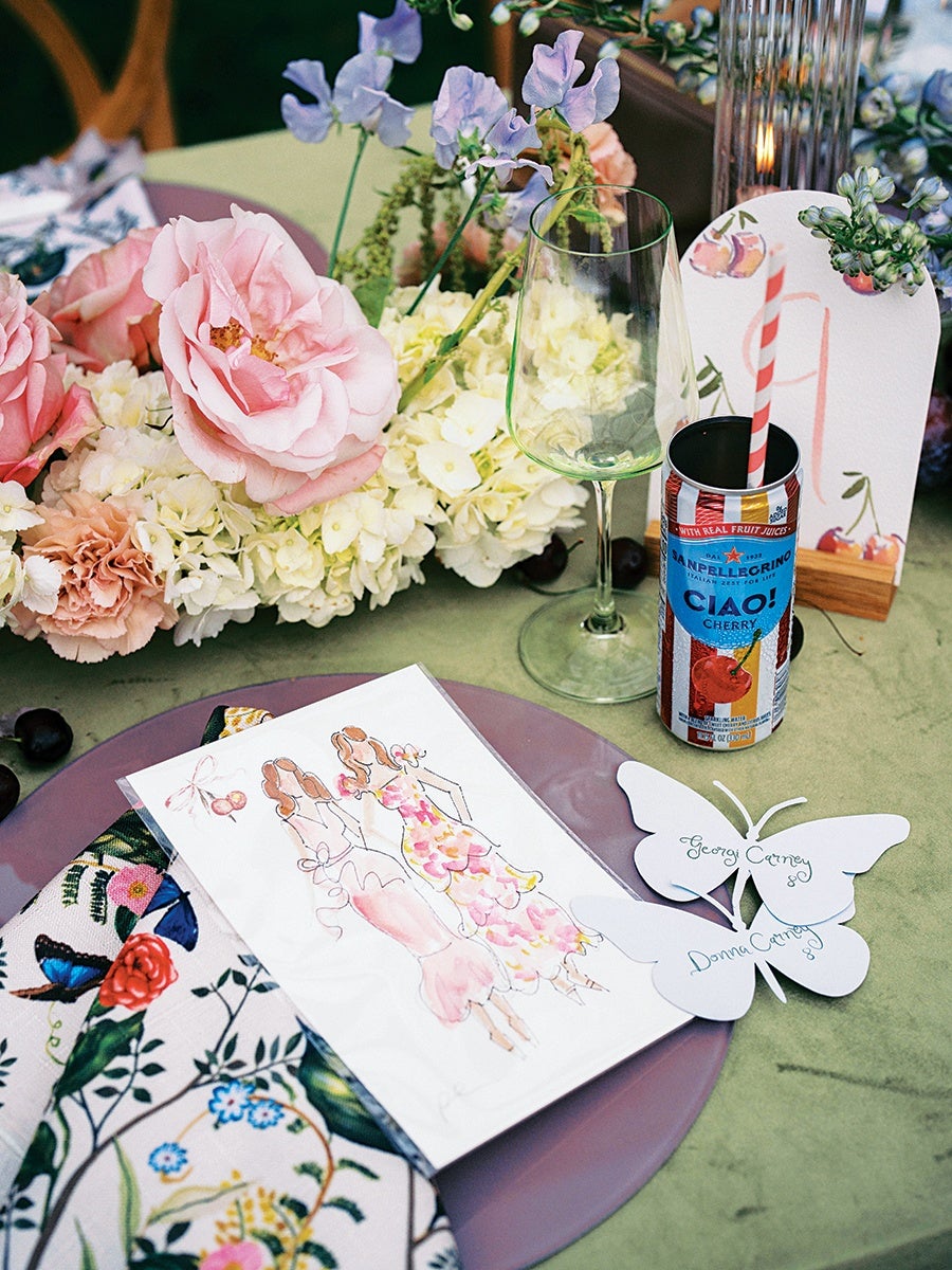 Table setting with a floral napkin, a purple plate, and a card featuring an illustration of two women in dresses. Two white butterfly-shaped place cards with the names "Georgi Carney" and "Donna Carney" are placed on the plate. A can of Sanpellegrino Ciao! Cherry drink with a striped straw is next to an empty wine glass. The background includes a floral centerpiece with pink roses, white hydrangeas, and purple flowers on a green tablecloth.