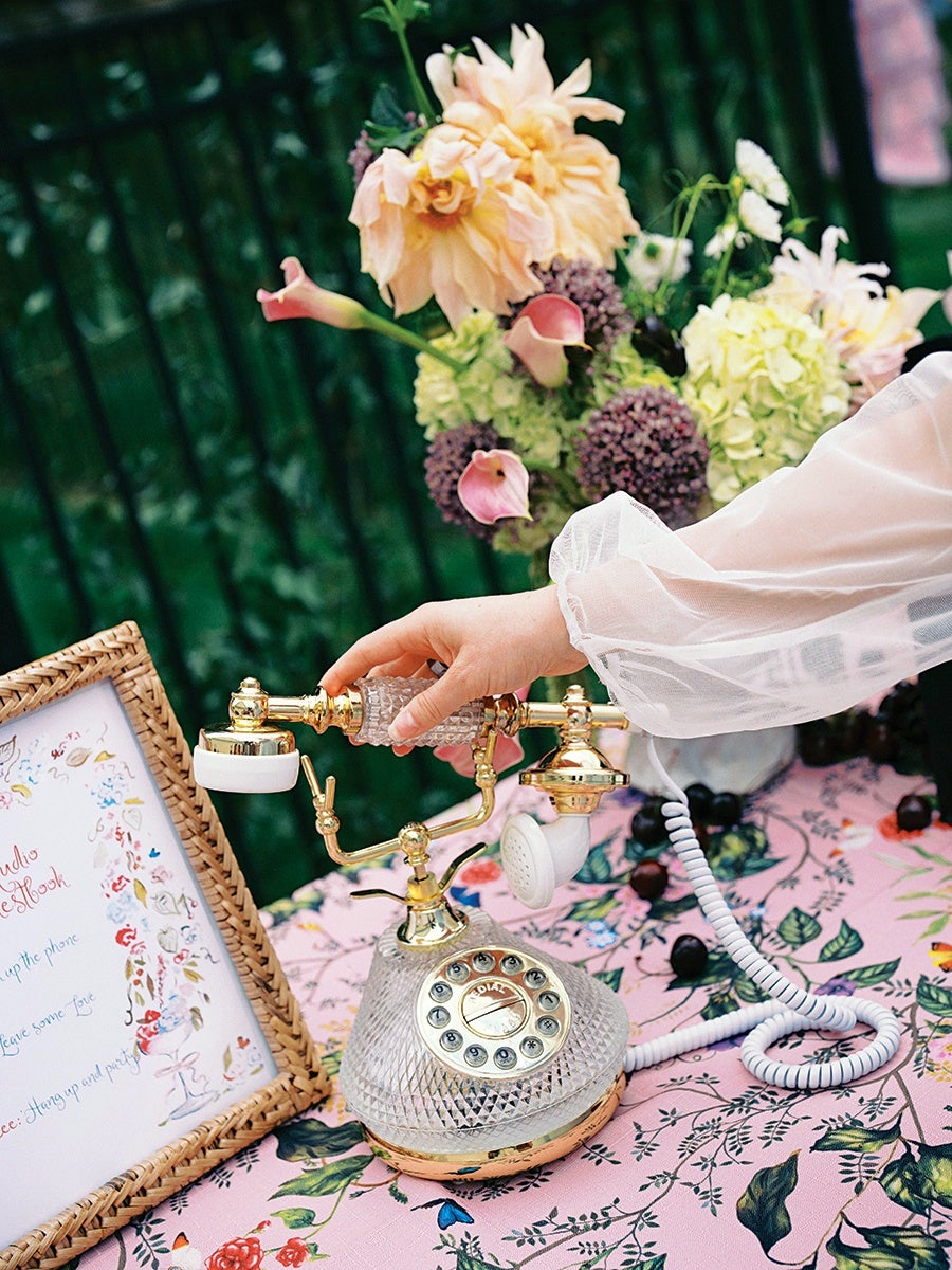 A vintage-style rotary telephone with a clear, textured glass body and gold accents sits on a pink floral tablecloth. A hand with a sheer white sleeve is lifting the handset. Behind the phone, there is a bouquet of flowers including pale peach, purple, and white blooms. To the left, a wicker-framed sign with colorful floral illustrations and cursive writing is partially visible.