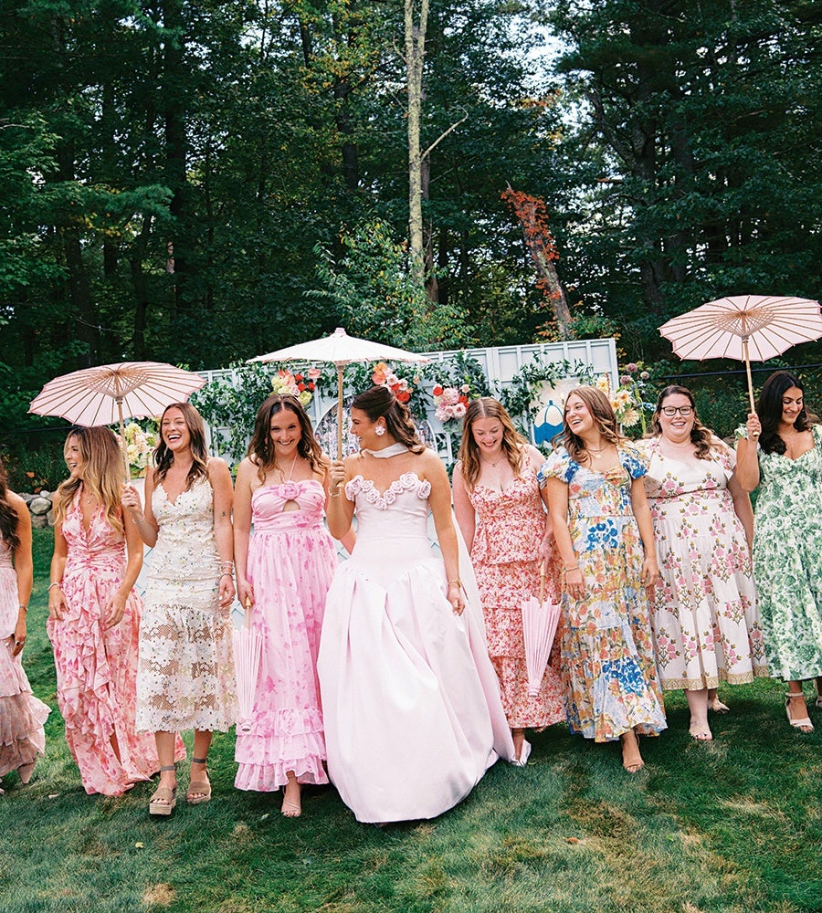 A group of women standing on grass in a garden setting, dressed in colorful floral and pastel dresses. The central woman wears a white gown with floral details on the bodice and holds a white parasol. Several women hold pink parasols, and there is a backdrop decorated with flowers and greenery behind them. Tall trees and dense foliage form the background.