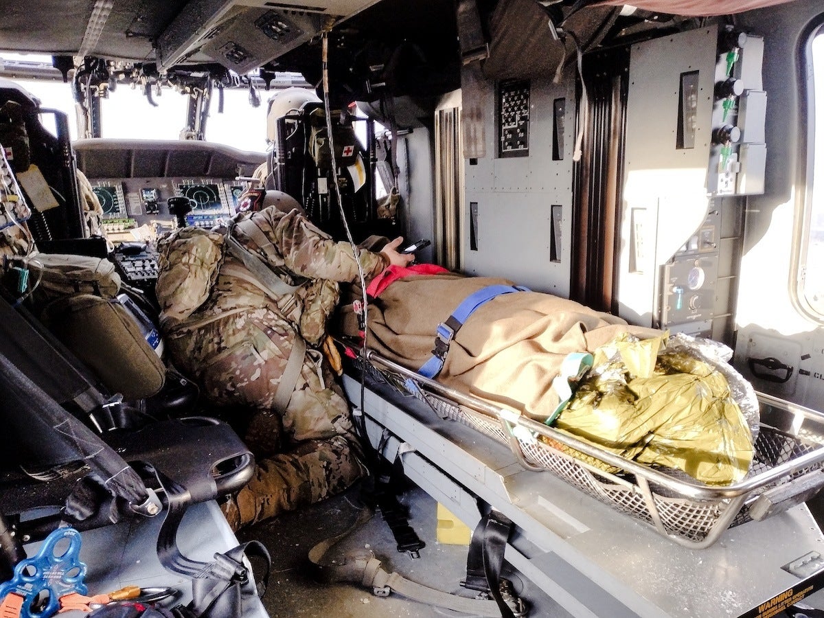 A soldier in camouflage uniform kneels inside a military helicopter, attending to a person secured on a stretcher with a blue strap. The person on the stretcher is covered with a brown blanket and a metallic emergency blanket. The helicopter cockpit with various controls and instruments is visible in the background.