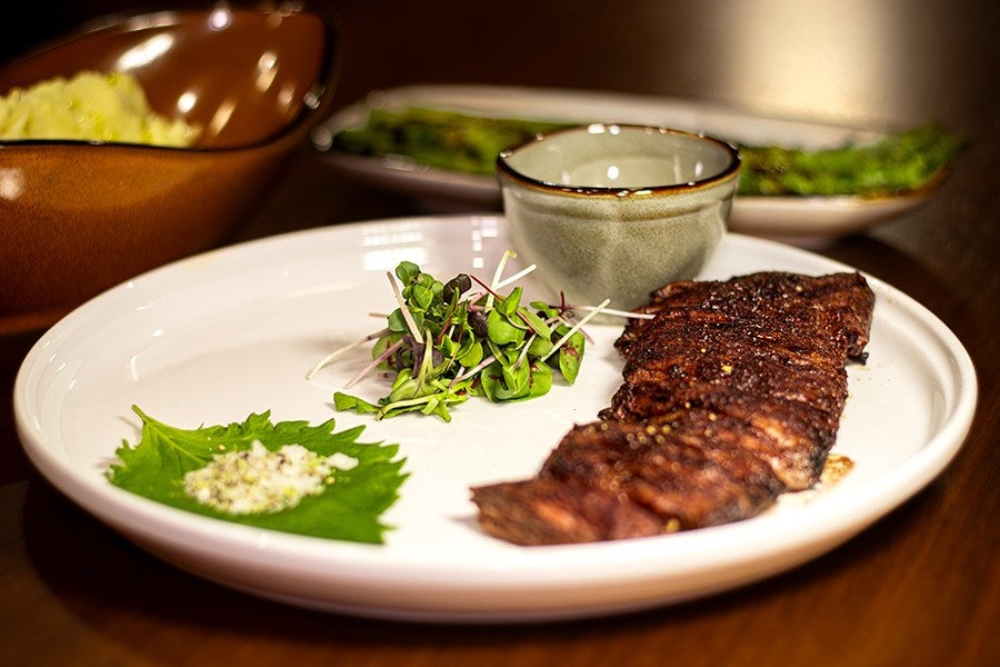 Grilled steak served on a white plate with a small pile of microgreens, a green leaf topped with seasoning, and a small ceramic bowl. In the background, there is a dish of grilled asparagus and a brown bowl containing mashed potatoes.