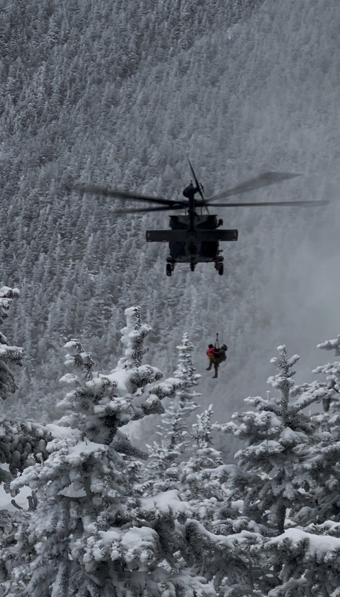 A helicopter hovers above a snowy forest, with two people being hoisted up by a rescue cable. The trees below are covered in thick snow, and the background shows a dense, snow-covered forest. The scene suggests a mountain rescue operation in a wintery environment.