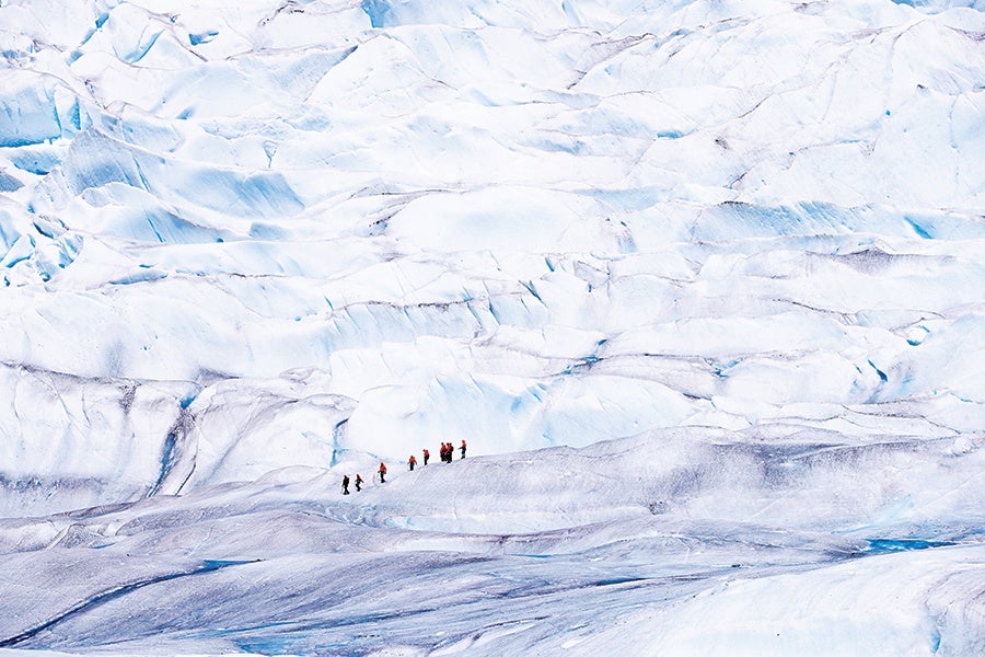 A group of hikers wearing red jackets trekking across a vast, icy glacier with textured, snow-covered surfaces and deep blue crevices.