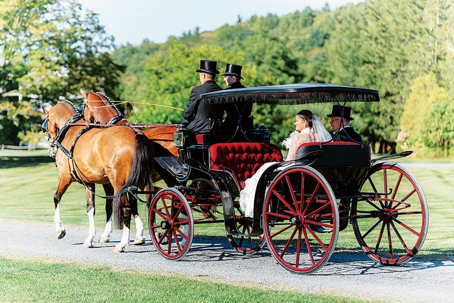 A horse-drawn carriage with red wheels and black body carries a bride in a white dress and veil, accompanied by a man in a black suit and top hat. Two men in black suits and top hats are seated at the front, driving two brown horses. The scene is set on a gravel path with green grass and trees in the background.