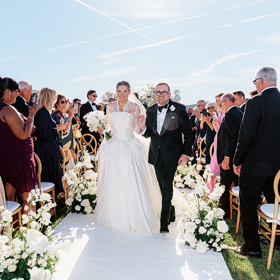 Bride and groom walking down a white aisle outdoors, surrounded by guests seated on wooden chairs. The bride wears a white gown with lace sleeves and holds a bouquet of white flowers, while the groom wears a black tuxedo with a bow tie and glasses. The aisle is decorated with white floral arrangements, and the sky is clear with visible contrails.