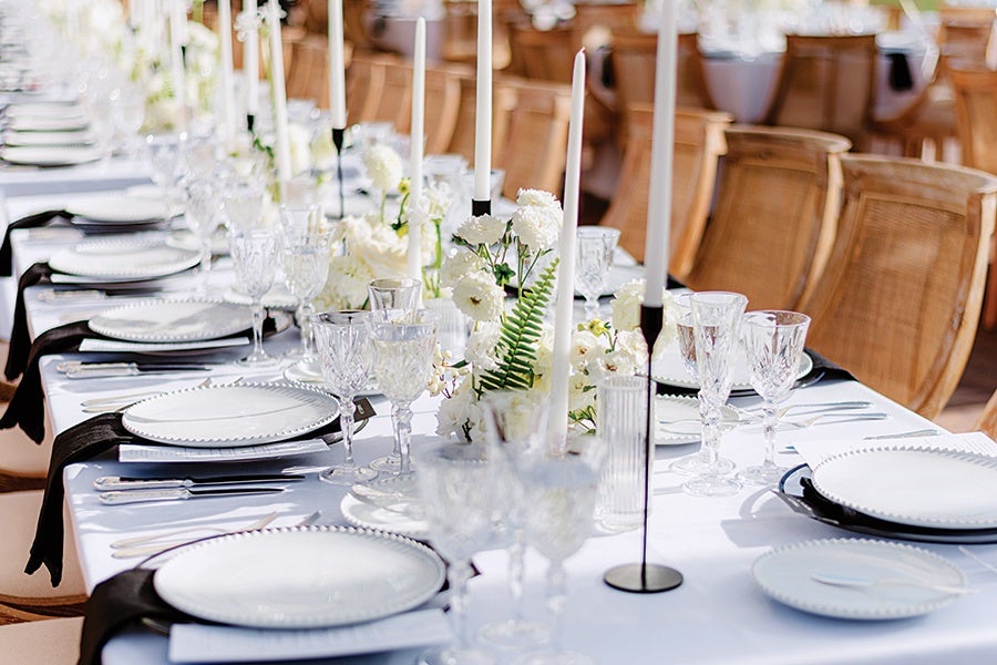 A long dining table set with white plates featuring a beaded edge, crystal glassware, and black cloth napkins. Tall white taper candles in black candle holders and white floral arrangements with greenery run down the center of the table. Wooden chairs with woven backs surround the table. The setting is bright and elegant.