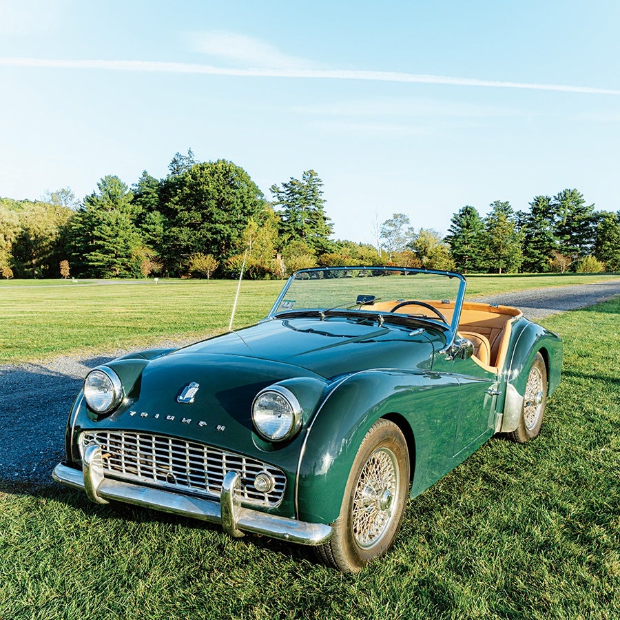 Dark green vintage Triumph convertible with tan interior, wire-spoke wheels, and chrome front bumper, parked on grass with trees and a clear sky in the background.