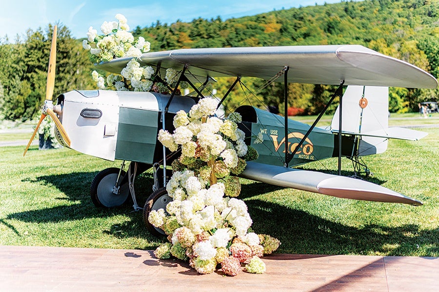 A vintage-style biplane model decorated with clusters of white and light pink hydrangea flowers, positioned on a grassy area with trees and hills in the background. The plane has a wooden propeller and the text "Vigo Motor Oil" on its side.