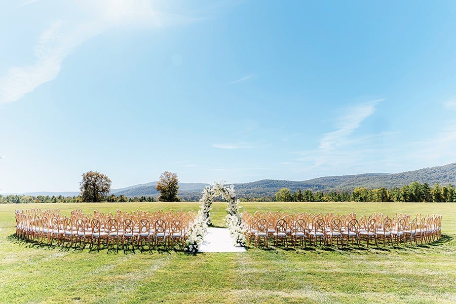 A circular outdoor wedding ceremony setup on a grassy field with wooden chairs arranged in a wide ring around a white aisle. At the center of the aisle is a floral arch decorated with white flowers. The background features distant trees and rolling hills under a clear blue sky.