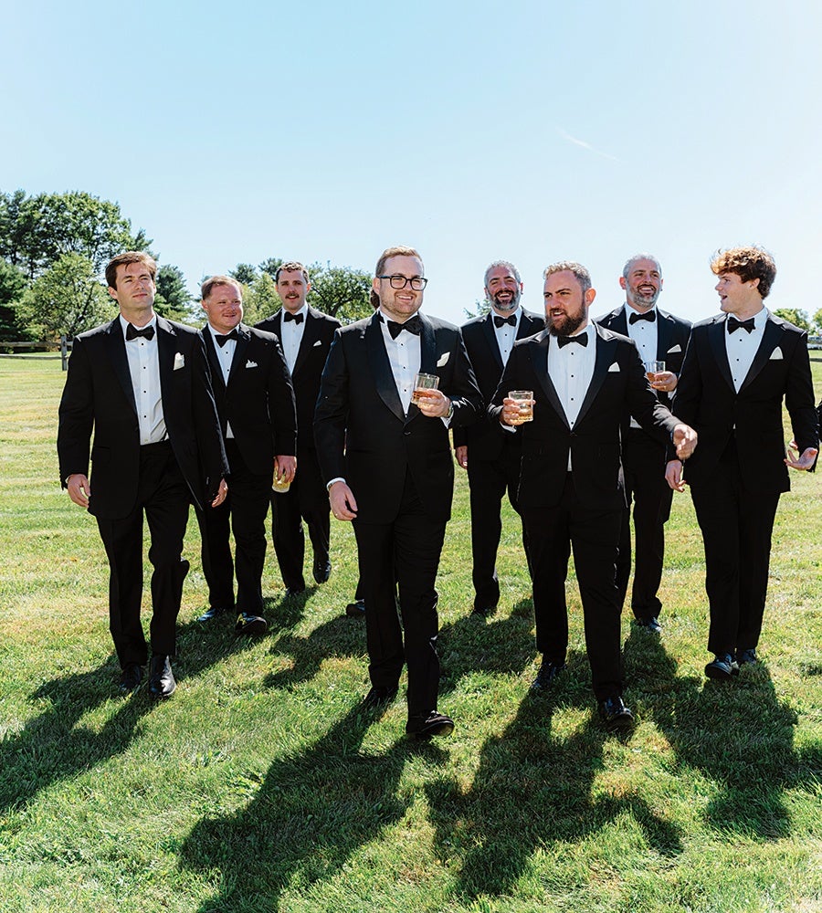 Eight men dressed in black tuxedos with bow ties are walking on a grassy field under a clear blue sky. Four of them in the front are holding glasses with drinks, and they appear to be smiling and engaged in conversation. Trees and a fence are visible in the background.