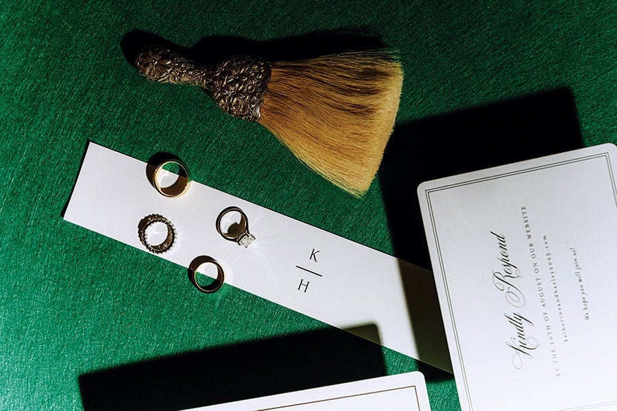 Four rings, including a diamond engagement ring and three gold bands, are displayed on a white card with the initials "K / H" printed on it, placed on a green surface. Nearby is a decorative brush with a carved handle and light brown bristles, along with a white card featuring elegant script text.