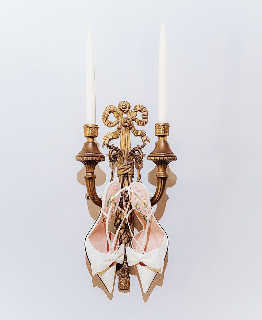 White pointed-toe high heels with large bows on the front, hanging from an ornate gold wall sconce with two white taper candles. The shoes have pink insoles with floral designs and the Valentino logo.