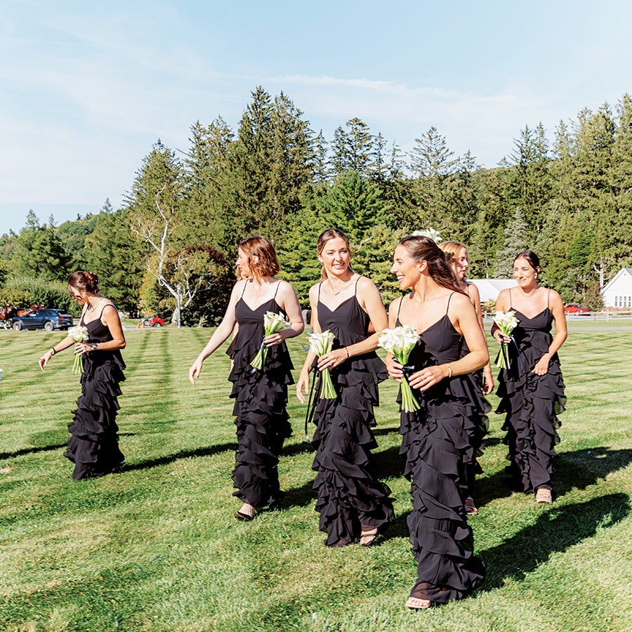 Six women wearing matching black ruffled dresses are walking on a grassy field, each holding a small bouquet of white flowers. The background features tall green trees and a clear blue sky.