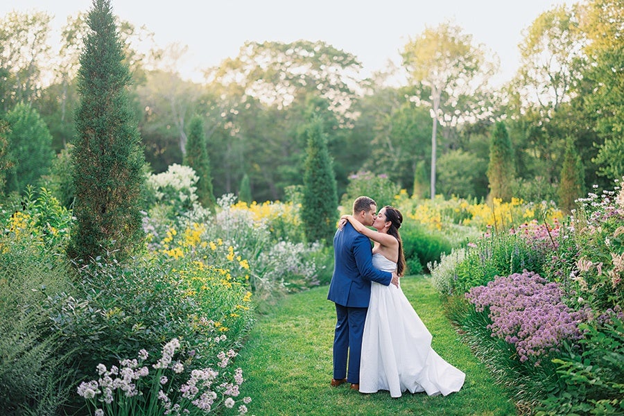 A couple embracing and kissing in a garden pathway surrounded by lush greenery and colorful flowers, with the woman wearing a white wedding dress and the man in a blue suit.