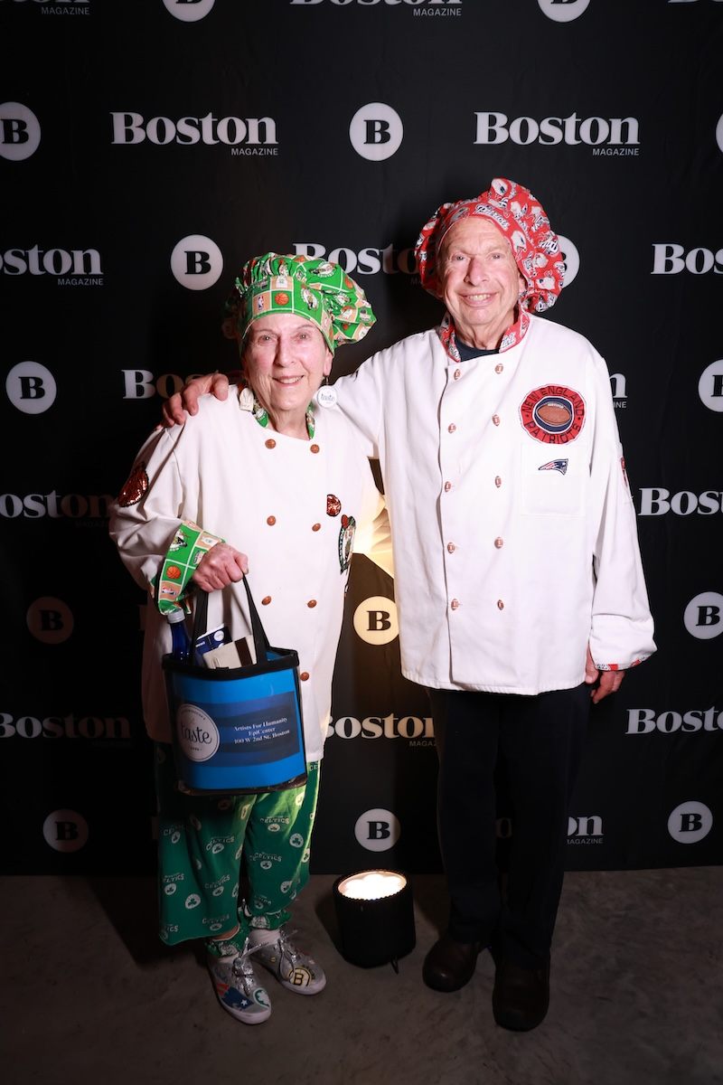 Two older adults wearing white chef jackets and colorful chef hats stand in front of a black backdrop with "Boston Magazine" logos. The person on the left wears a green hat and pants with sports-themed prints and holds a blue tote bag. The person on the right wears a red hat with a sports logo patch on the jacket. Both are smiling.