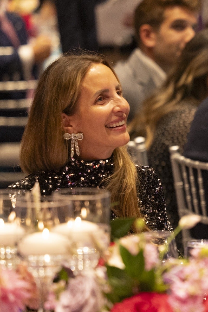 A woman with long brown hair is smiling and looking to her right. She is wearing a black sequined top and large, sparkly bow-shaped earrings. In the foreground, there are blurred candles and colorful flowers, suggesting a formal or festive event. Other people are visible in the background, seated and dressed formally.