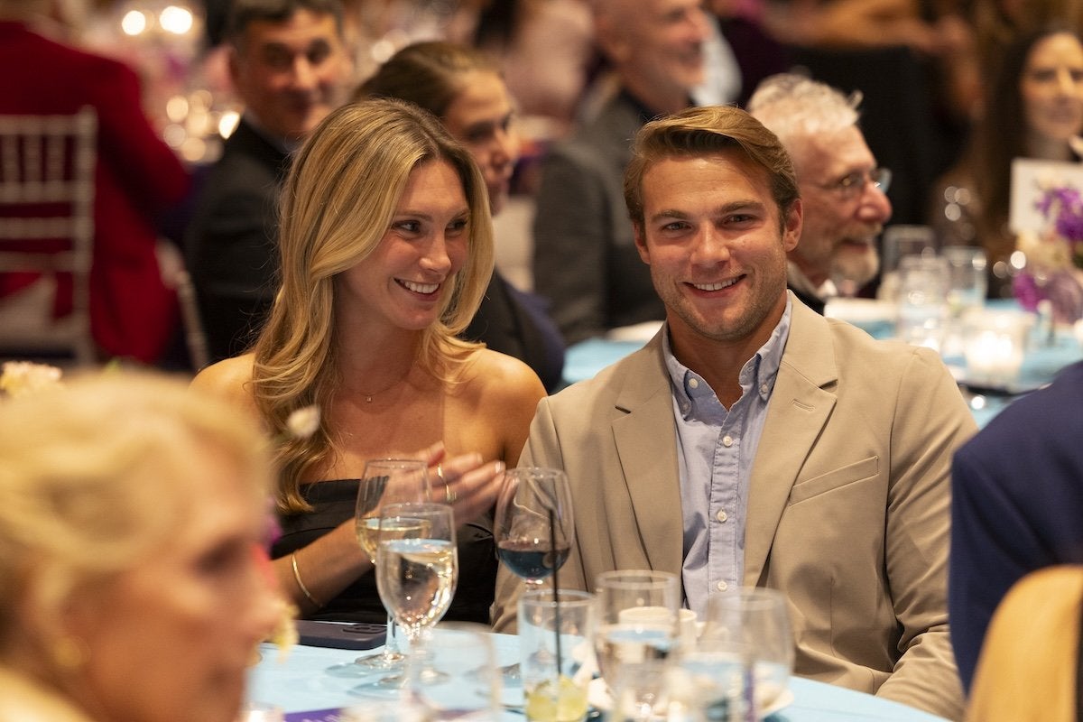 A smiling man and woman sitting at a table during a formal event. The woman has long blonde hair and is wearing a strapless black dress, while the man is wearing a beige blazer over a light blue shirt. The table is covered with a light blue tablecloth and has several glasses of water and wine. Other people are visible in the blurred background.