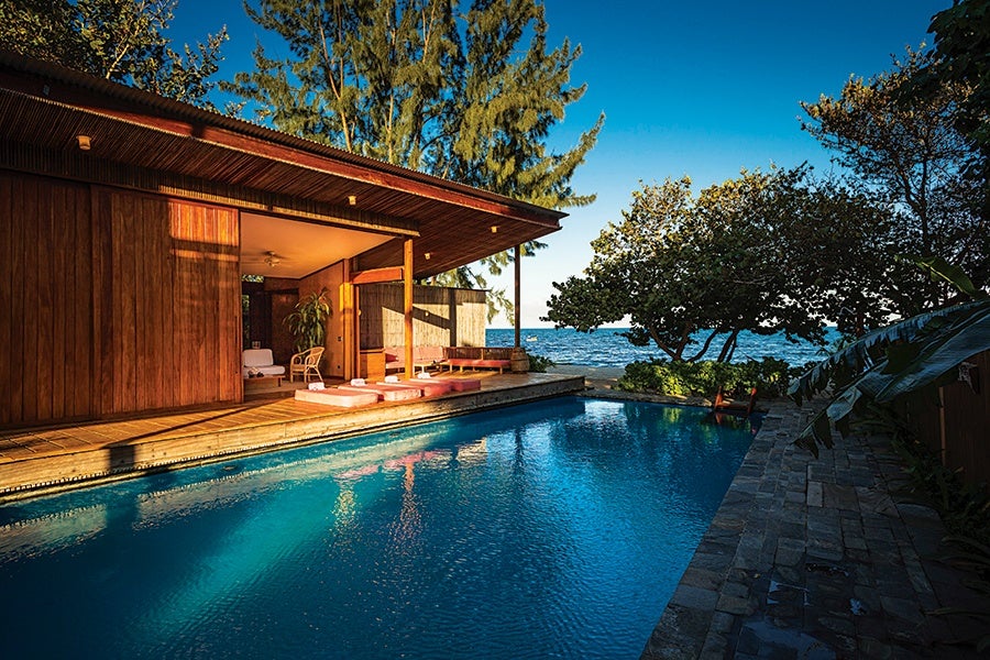 A modern wooden house with an open patio featuring lounge chairs and cushions next to a rectangular swimming pool. The pool area is surrounded by stone tiles and lush greenery, with trees and the ocean visible in the background under a clear blue sky.