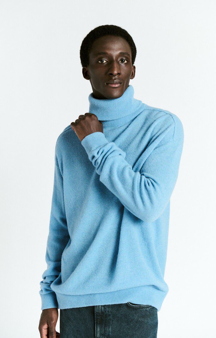 A man wearing a light blue turtleneck sweater and dark jeans, standing against a plain white background.