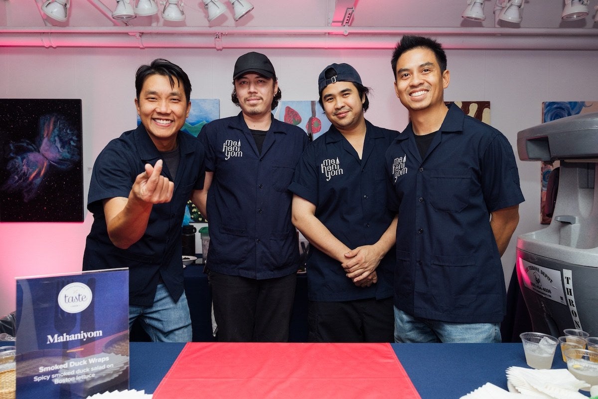 Four men wearing matching dark blue shirts with the logo "mahaniyom" stand behind a table covered with a blue and red tablecloth. The man on the far left is smiling and making a finger heart gesture. On the table, there is a sign that reads "Mahaniyom" and describes "Smoked Duck Wraps" with spicy smoked duck salad on Boston lettuce. The background features colorful paintings and overhead lighting.
