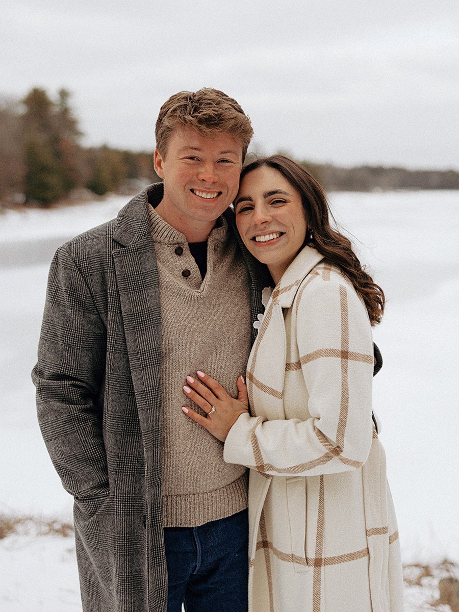 A smiling couple standing close together outdoors in a snowy landscape. The man is wearing a gray plaid coat over a beige sweater and blue jeans, while the woman is wearing a white coat with beige plaid patterns. The woman has her hand resting on the man's chest, showing a ring on her finger. Both have dark hair, and the background features snow-covered ground and blurred trees.
