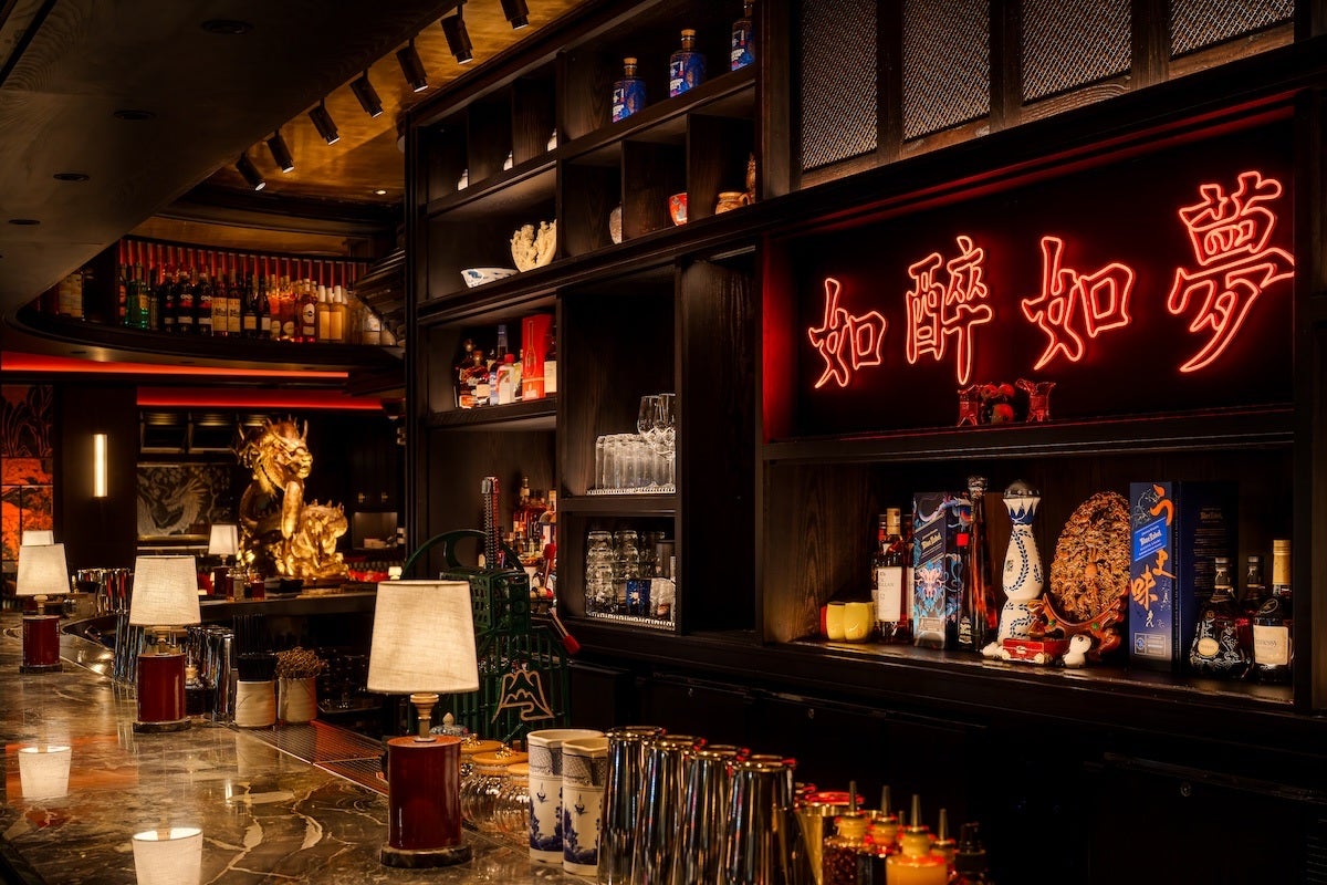 A dimly lit bar with a polished marble countertop featuring several small lamps with beige shades. Behind the bar, dark wooden shelves hold various bottles of liquor, glassware, and decorative items. A prominent red neon sign with Chinese characters is mounted on the wall above the shelves. In the background, a golden dragon statue adds an ornate touch to the ambiance. The overall atmosphere is warm and inviting with an Asian-inspired theme.