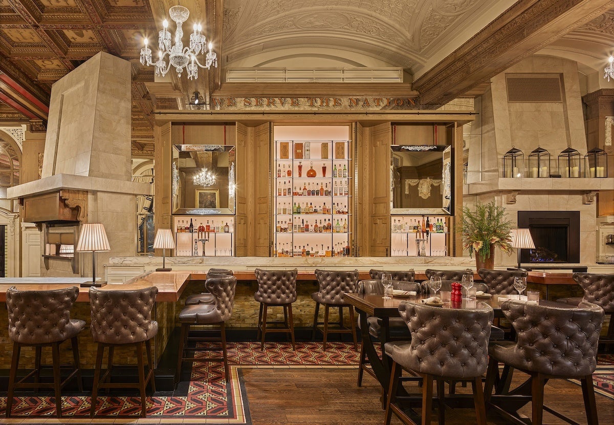 Luxurious bar area with tufted leather chairs arranged around wooden tables and a marble countertop. The back wall features a well-lit display of various liquor bottles framed by wooden panels and mirrors. Elegant chandeliers hang from a detailed ceiling, and the space is accented with table lamps, a fireplace, and decorative plants. The floor combines patterned carpet and wood.