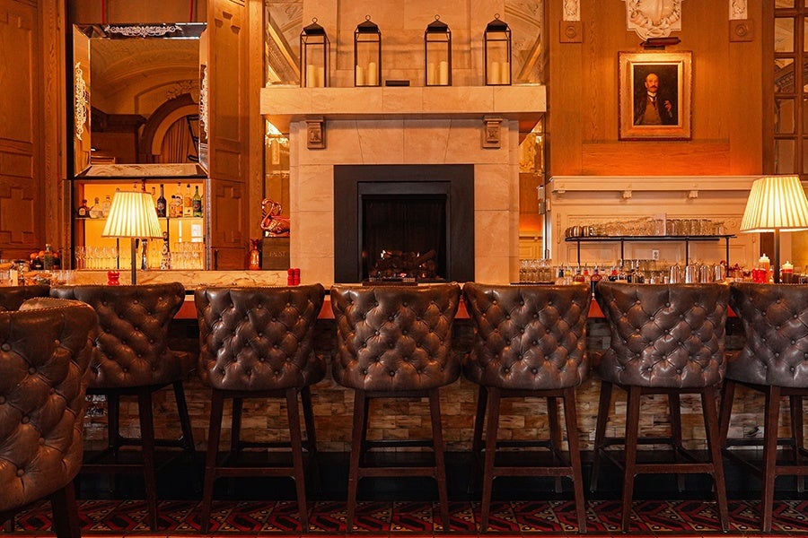 A row of six dark brown tufted leather bar stools lined up in front of a bar counter. The bar features a stone fireplace with a black interior and four lantern-style candle holders on the mantel. Two table lamps with pleated shades are positioned on either side of the bar. The background includes wooden paneling, a large mirror, shelves with bottles and glasses, and a framed portrait on the wall. The floor has a patterned carpet with red and black designs.
