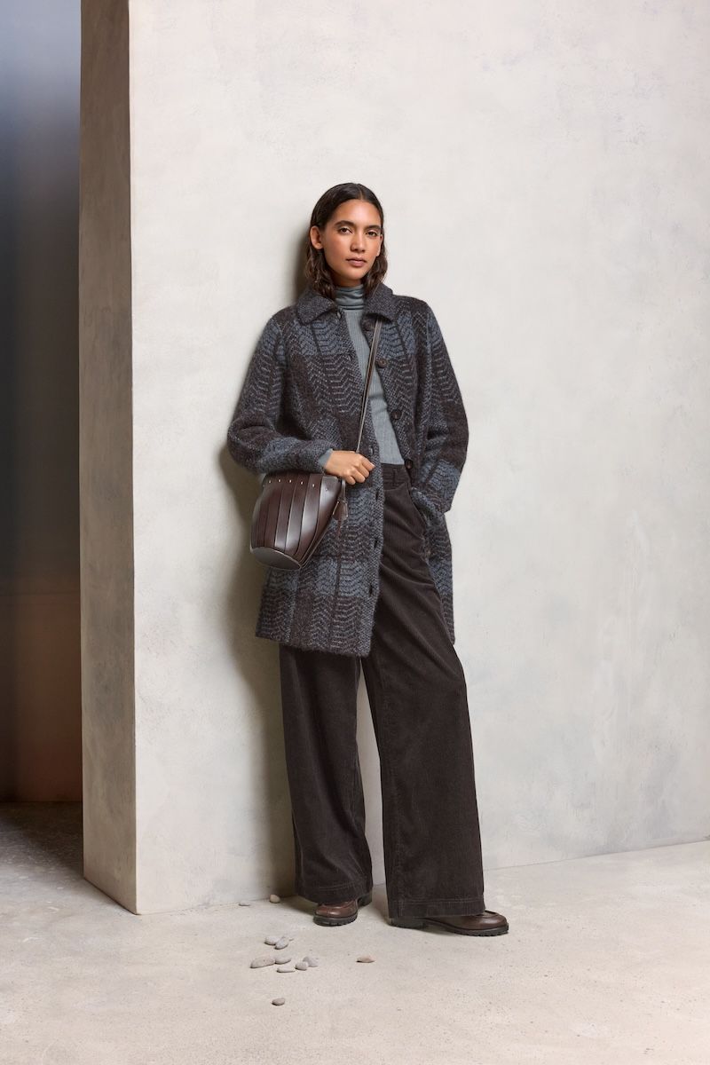 A woman wearing a long, textured dark gray cardigan over a light gray turtleneck sweater, paired with wide-leg black pants and dark brown shoes. She carries a dark brown, structured crossbody bag and stands against a light-colored textured wall with a few small stones scattered on the floor near her feet.