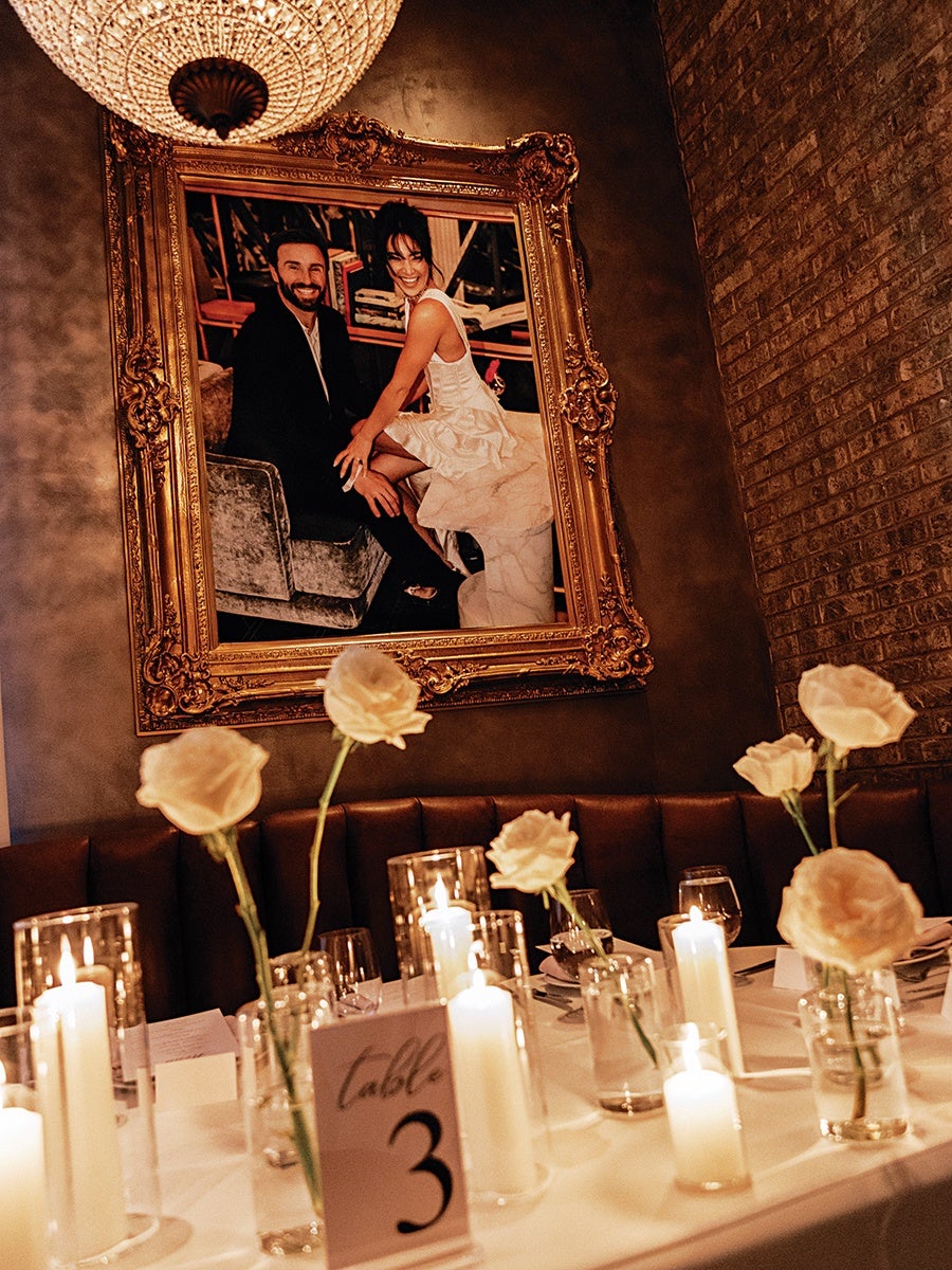 A warmly lit dining table set with white roses in glass vases and several lit white candles in glass holders. A table card labeled "Table 3" is visible. Behind the table is a brown leather bench against a wall with a large ornate gold-framed portrait of a smiling couple, the woman in a white dress and the man in a dark suit. The wall has a textured, dark finish with a brick section on the right side. A large, elegant chandelier hangs above.