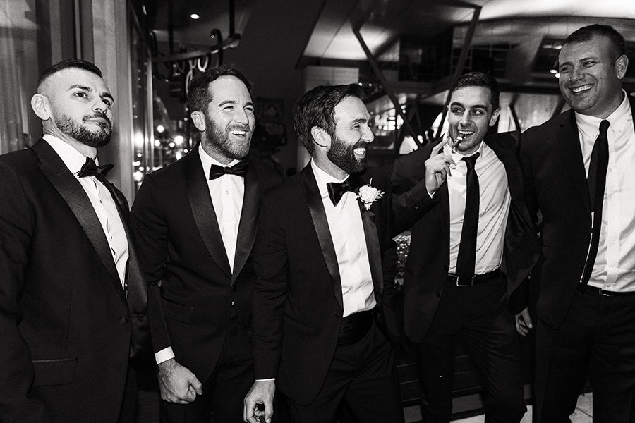 Five men dressed in formal black tuxedos and bow ties, standing closely together indoors, smiling and engaging with each other. One man in the center has a boutonniere on his lapel, suggesting he might be the groom. The setting appears to be a celebratory event, possibly a wedding.