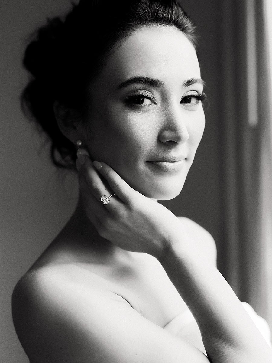 Black and white portrait of a woman with dark hair styled up, wearing a strapless top. She is gently touching her neck with one hand, showcasing a large, sparkling ring on her ring finger. Her expression is soft and serene, with subtle makeup highlighting her eyes and lips. The background is softly blurred, drawing focus to her face and hand.